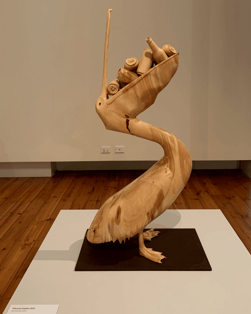 Wooden sculpture of a pelican with discarded bottles and drink cans in its mouth. 