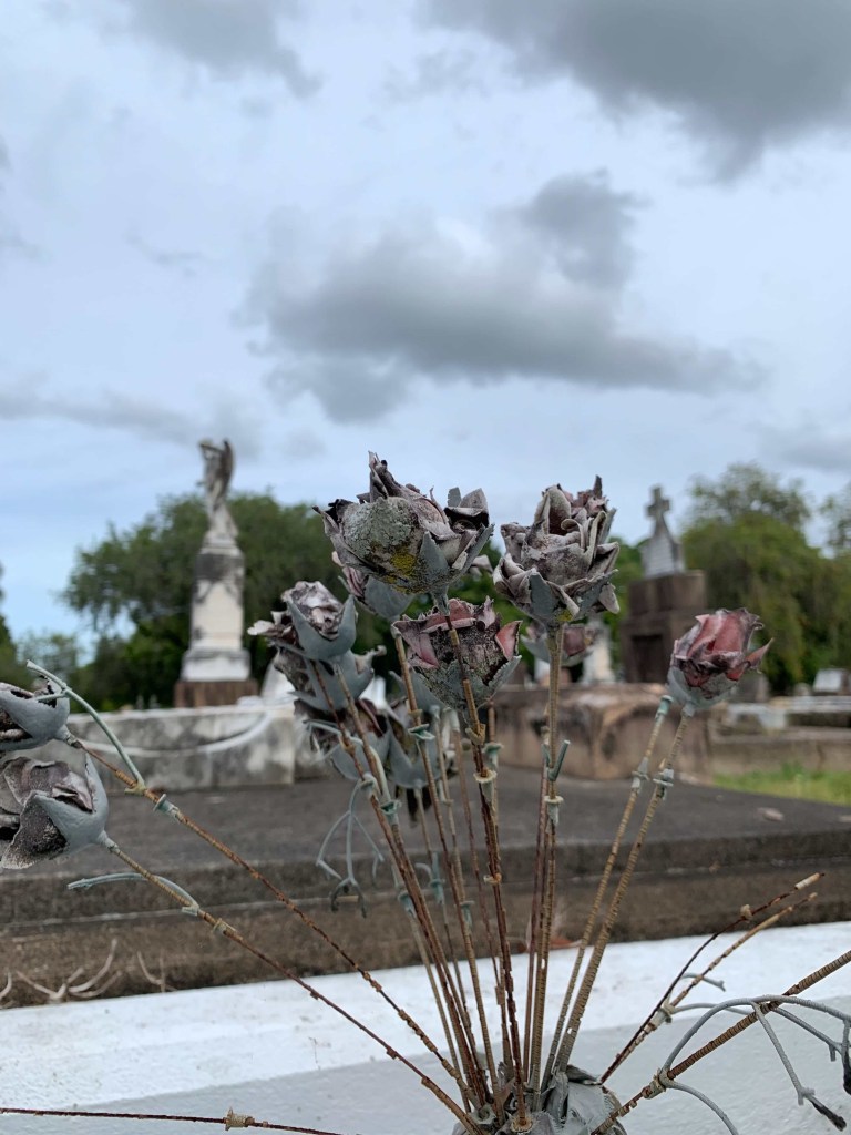 Close up photo of broken and crumbled leftovers of a bunch of plastic flowers on a grave stone. The wire stems of the flowers still remain, while most of the fake petals have broken up. 