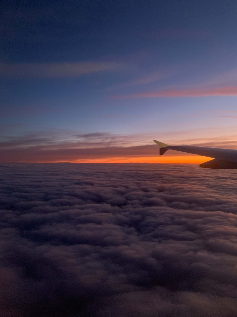 Photo of the sunset on a bed of clouds in the horizon, as seen from a plane. 
