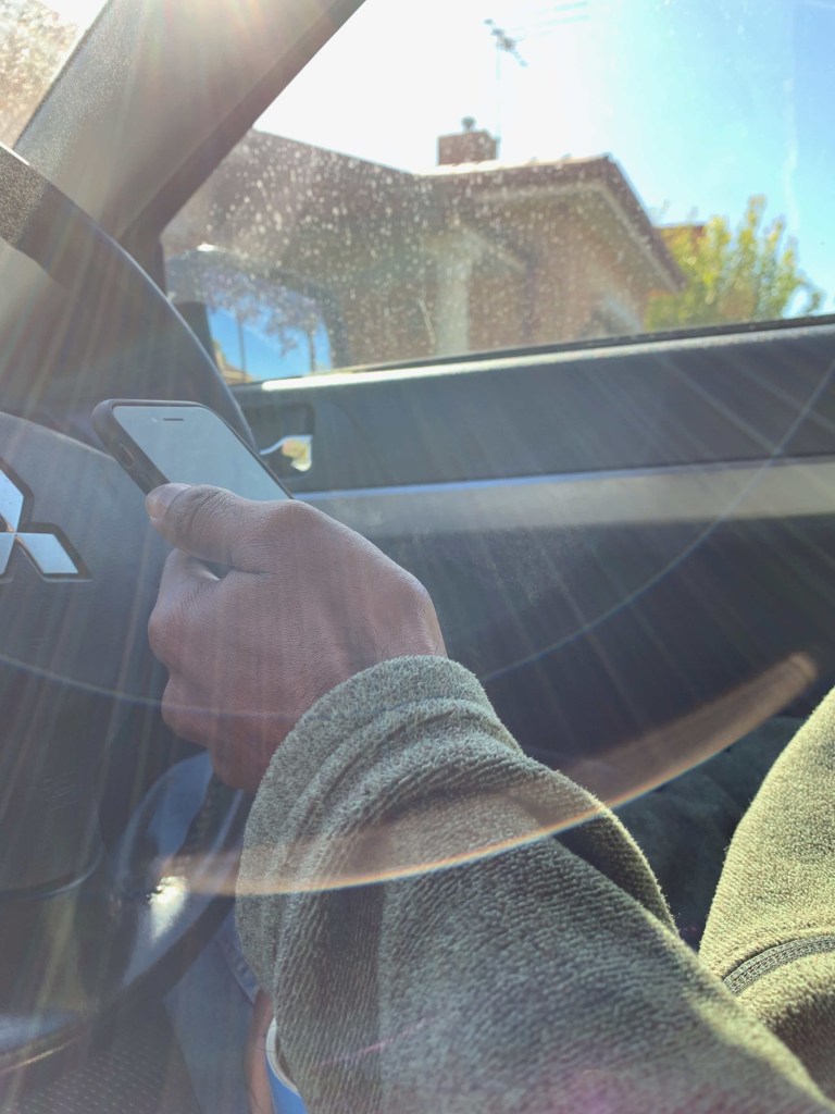 Photo of a person’s hand scrolling through the phone, while the person’s sat in the driver’s seat of a stationary car. The morning sun amines into the car, onto the person’s hand. 