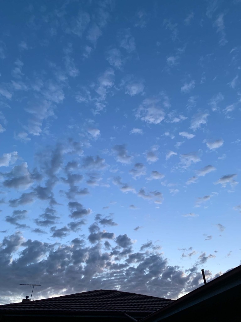 Photo of dark clouds in the sky, most of it clumping towards the background of the photo, over the roof of a house. With more clouds scattered in the foreground. 