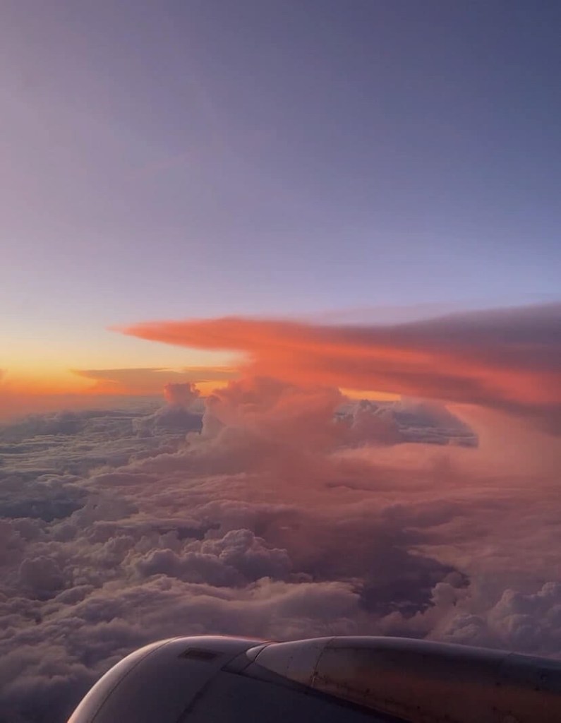 Photo of a big cloud at sunset, surrounded by and orange glow, as seen from the window seat of a plane. 