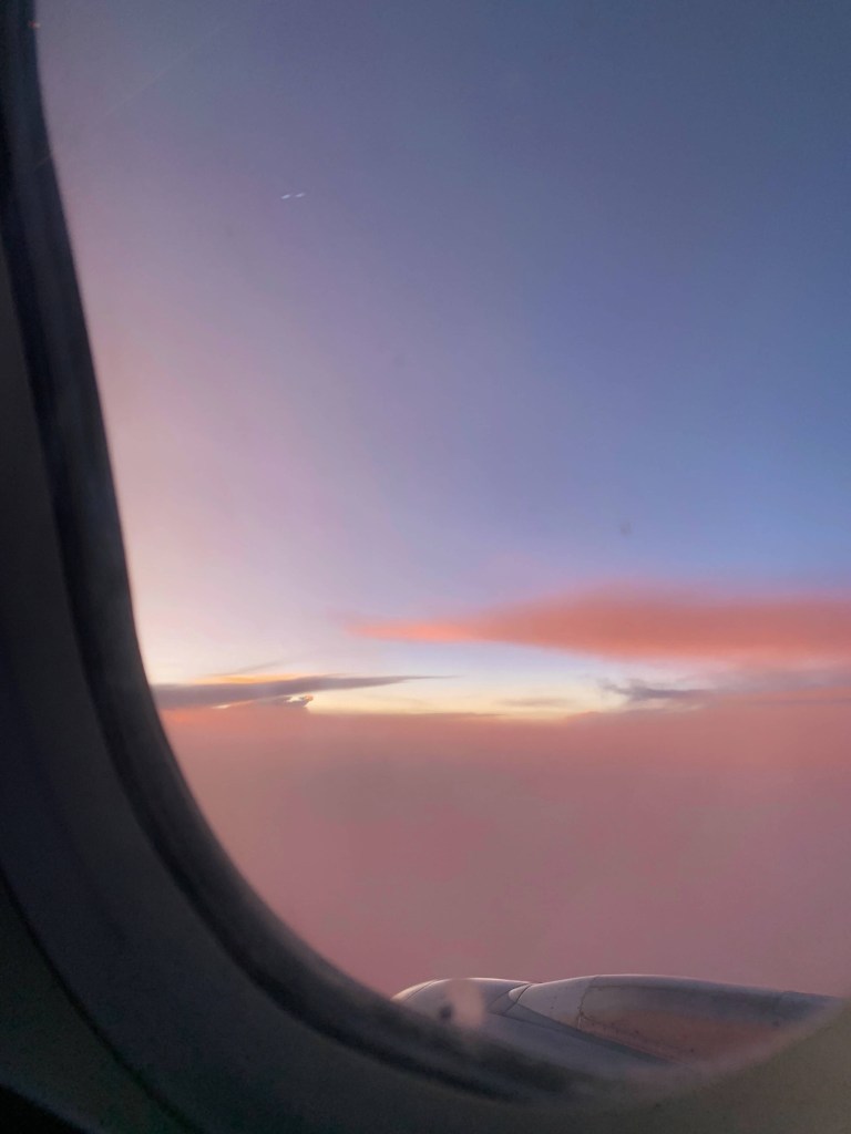 Photo of wispy, orange clouds at sunset, as seen through a plane’s window seat. 