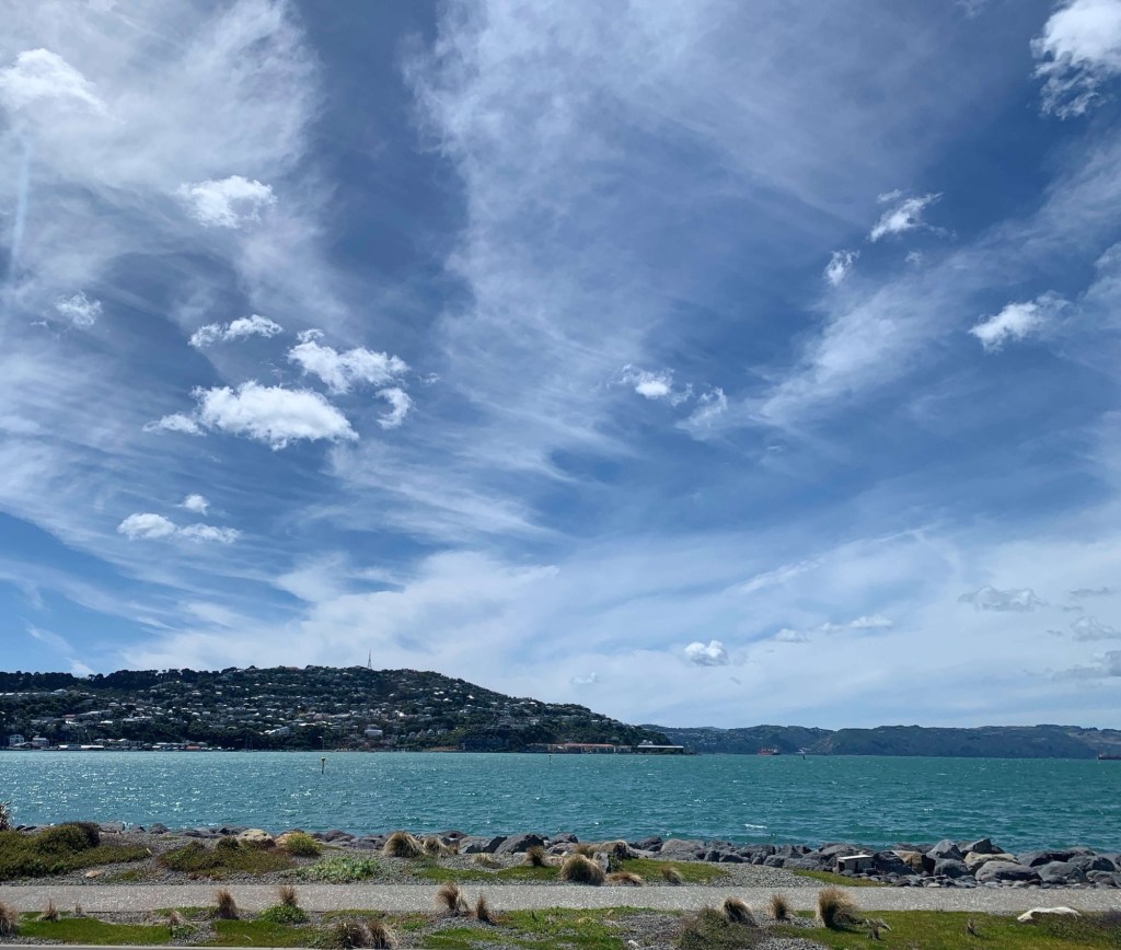 Photo of the ocean, as seen from the road with clouds spread out, appearing as if to move left to right. 