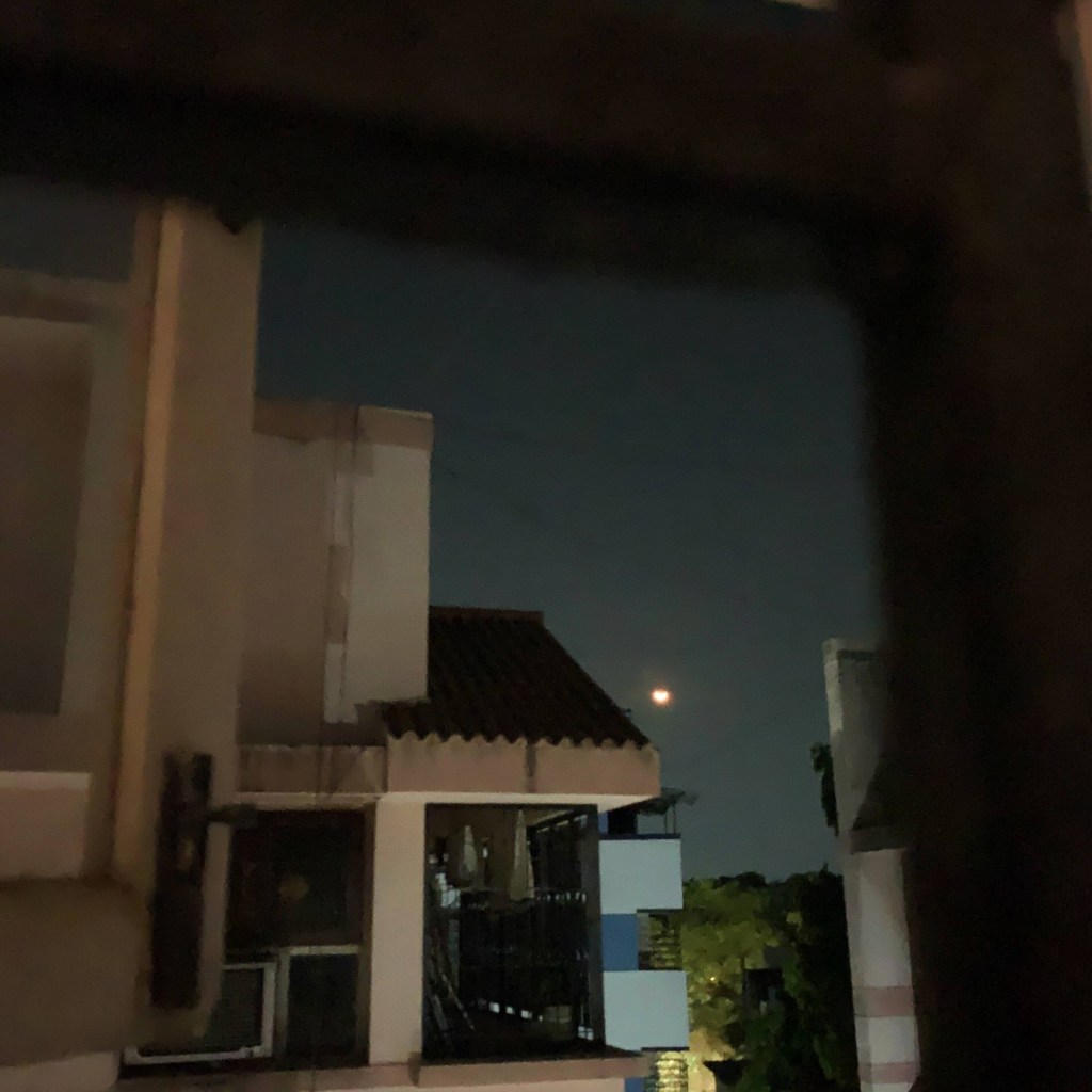 Photo of a half-moon, as seen from the window of a second floor building, with other buildings and facades visible. 