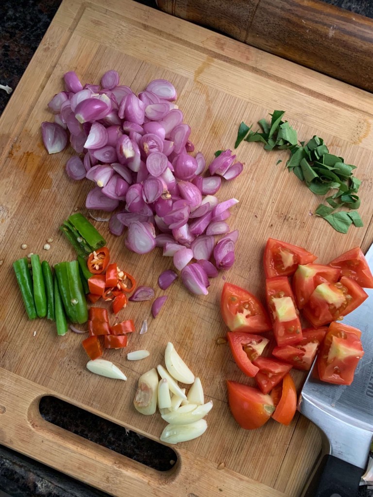 Close up photo of chopped onions, curry leaves, tomatoes, garlic, and green chillies on a wooden cutting board. 