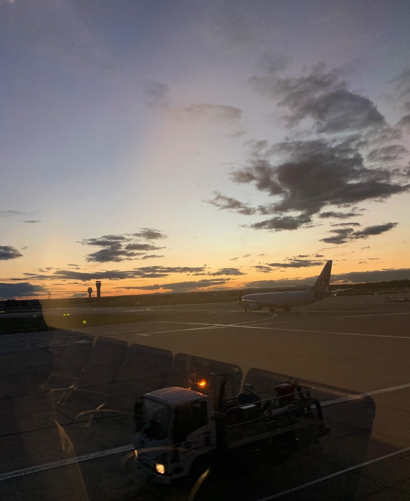 Photo of the sunset on the runway, as seen through the glass window of an airport. There’s an aircraft on the runway and a ground vehicle. The airport’s seating area is reflected on the glass. 