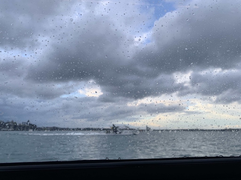 Photo of raindrops on a window of a ferry, with a view of dark clouds and the ocean.
