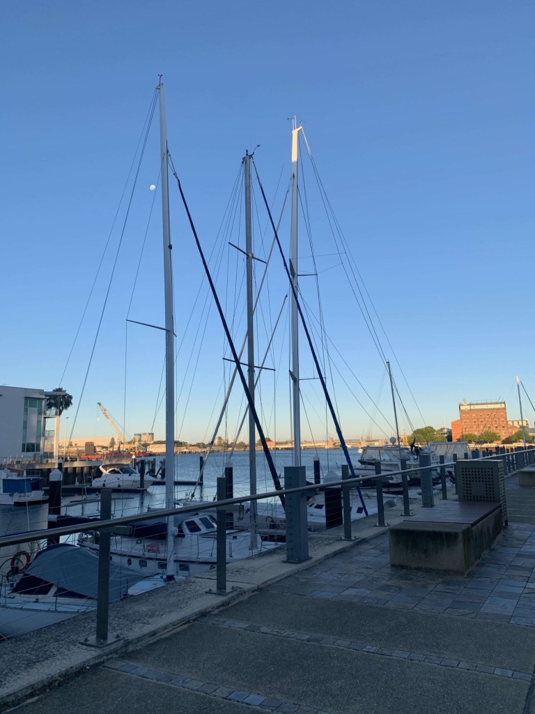 Photo of seven tall ships docked on the port at sunset, with the moon visible through the sails on one ship. 