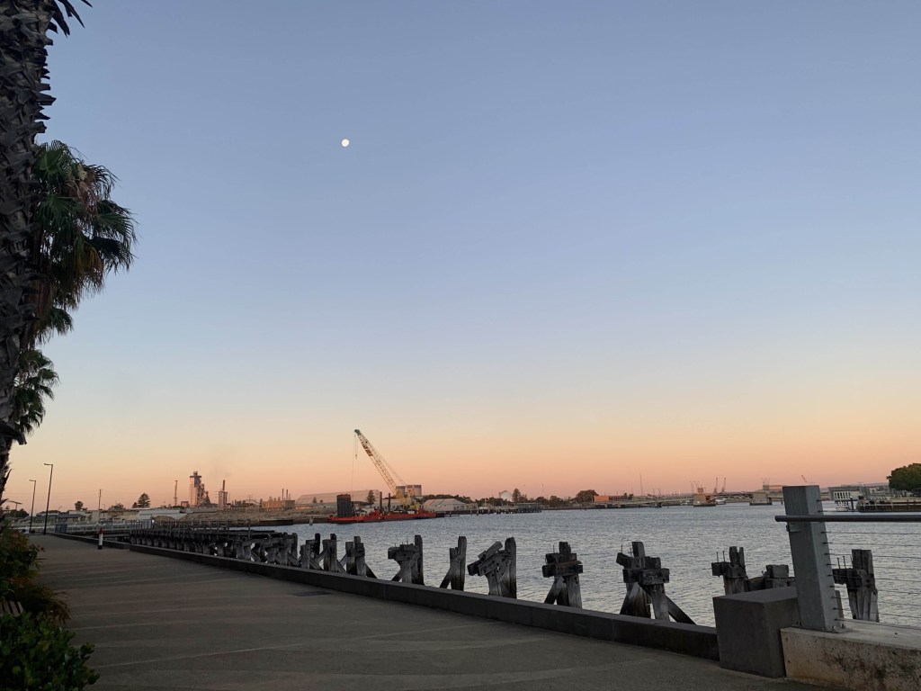 Photo of the sunset on the horizon, with the moon above and a crane in the distance. 