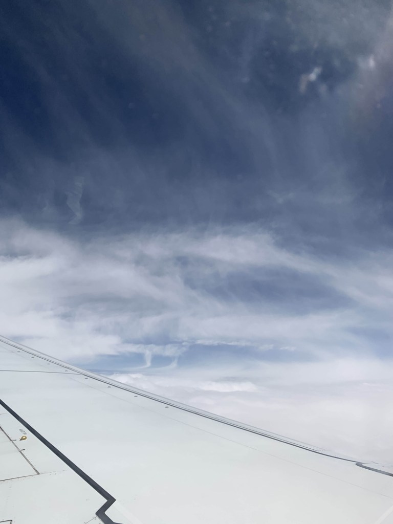 Photo of wispy clouds against a blue sky, as seen through the window of a plane. The plane’s wing is visible. 