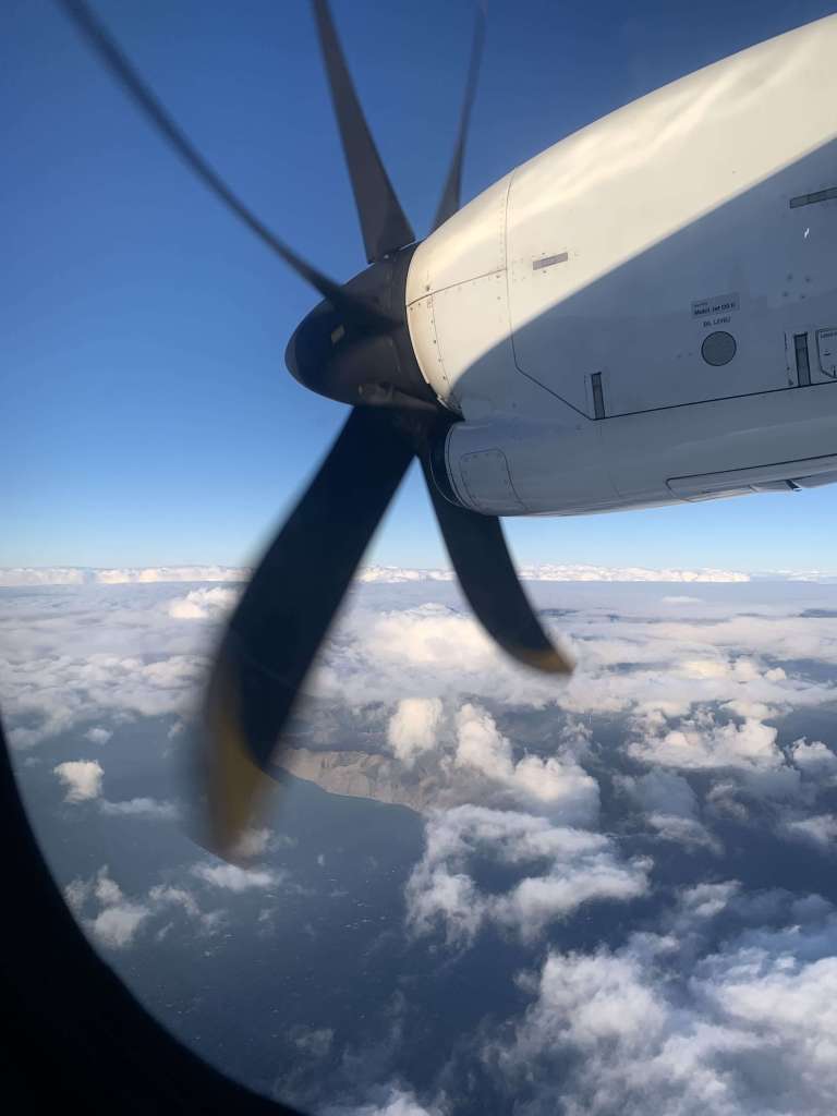 Photo of the coast of Wellington, New Zealand, in the distance, as seen from the window of a plane with a vibrant blue sky and scattered clouds.