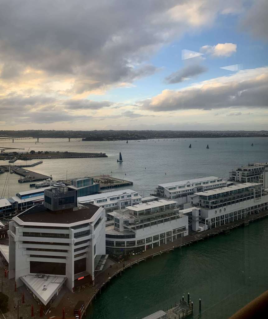 photo of light sunset over the Auckland city shoreline, as seen from the window of a tall building with boats on the water