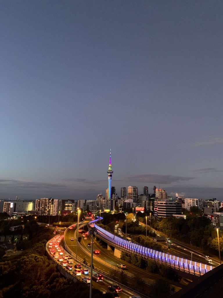 Photo of Auckland’s Sky Tower, lit up in multiple colours at sunset, along with the traffic on the motorway below. 