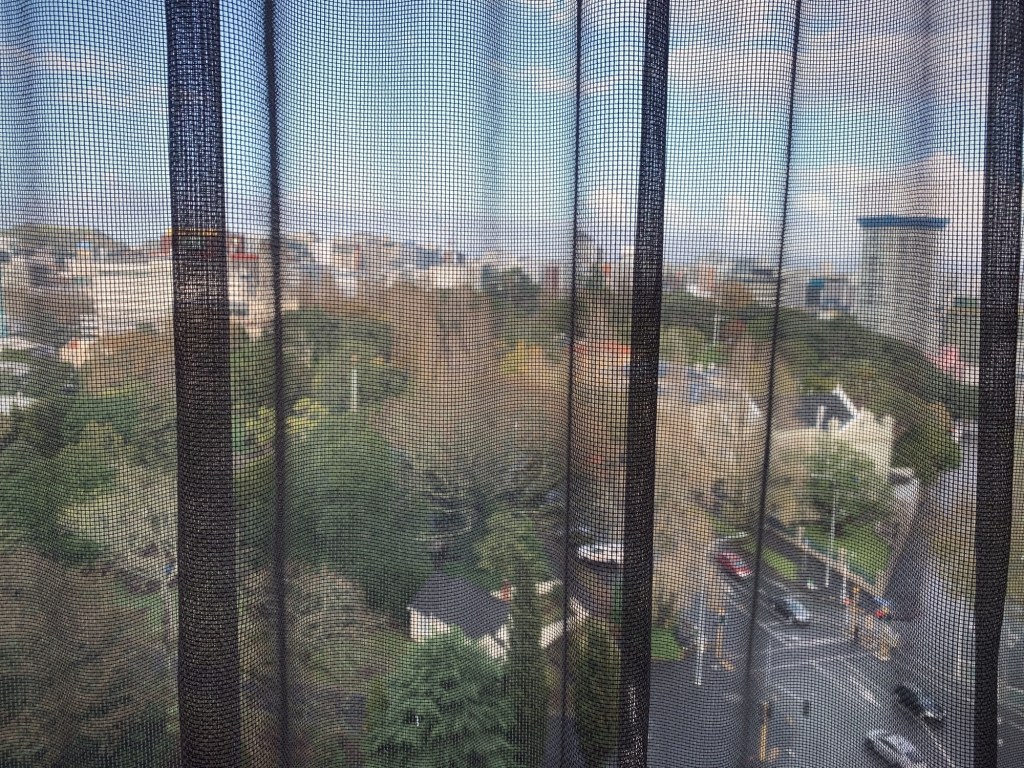 Photo of the Auckland city as seen through a lace-curtained window on a high floor of a hotel. 