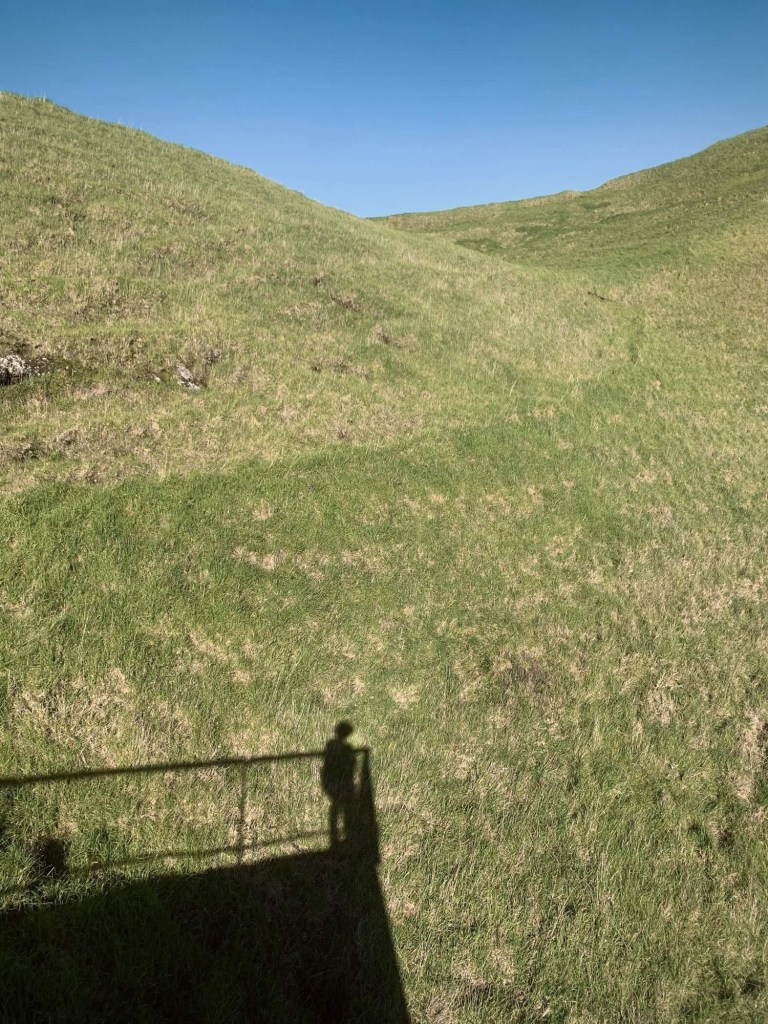Photo of the shadow of a person standing by the railings at top of a volcanic crater, looking down.