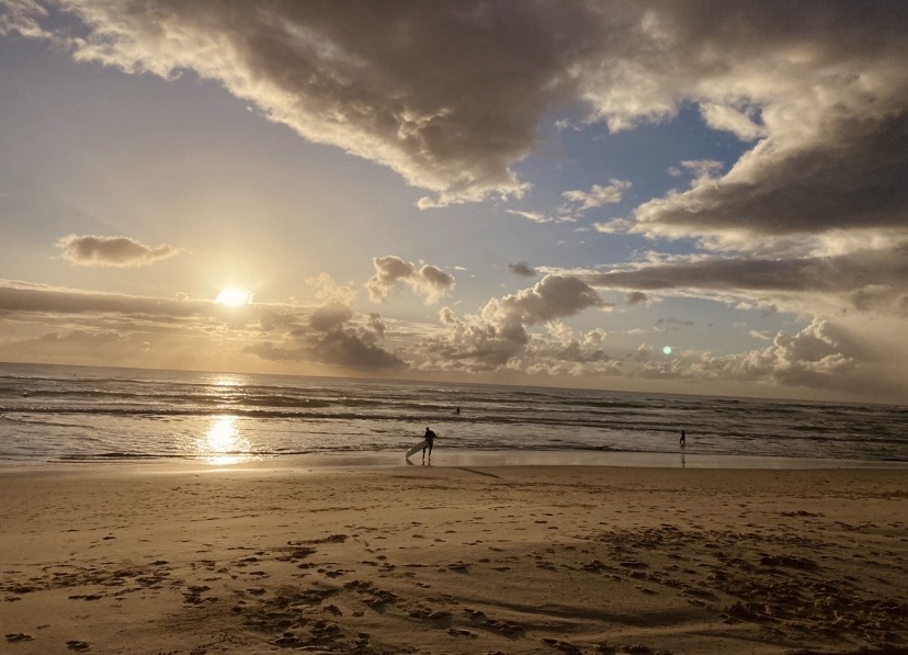 Photo of a beach and sunset and a lone surfer in the shore with a surfboard.