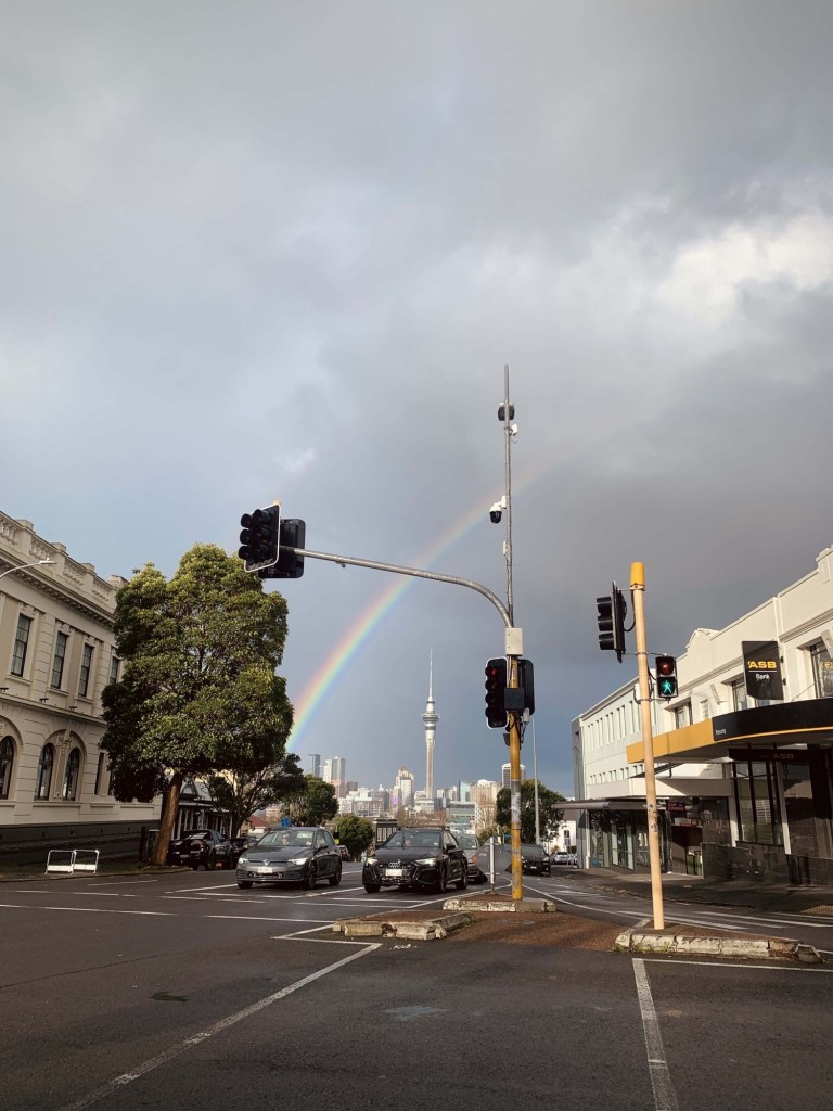 Photo of a rainbow over the Sky Tower of Auckland, as seen from a suburban street. 